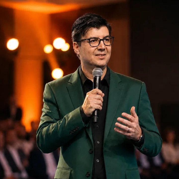 Johnis Pastori Jr. no palco, de terno verde e microfone, ensinando construções financiadas